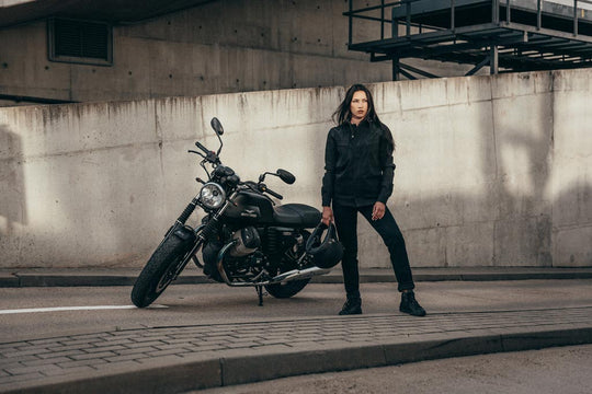 A woman rider in a stylish black leather jacket and jeans stands confidently next to her motorcycle, representing the modern female motorcyclist.