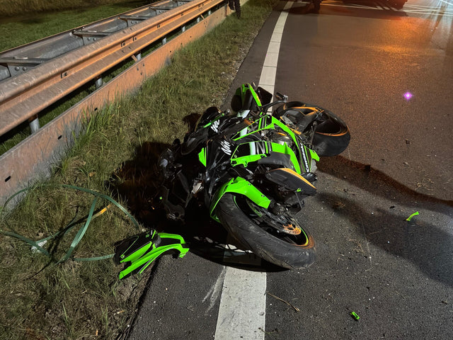 A heavily damaged green sport motorcycle lying on the roadside after a crash, near a highway guardrail at night with emergency lights in the background.