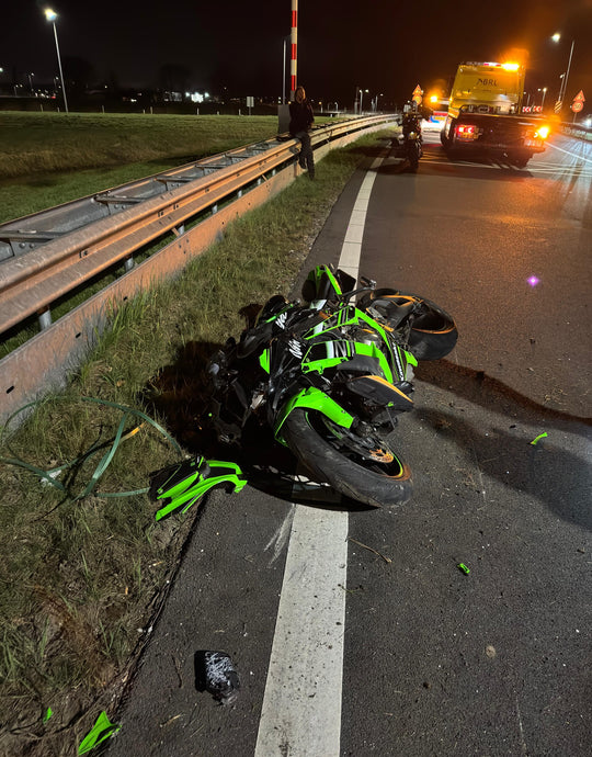 A heavily damaged green sport motorcycle lying on the roadside after a crash, near a highway guardrail at night with emergency lights in the background.